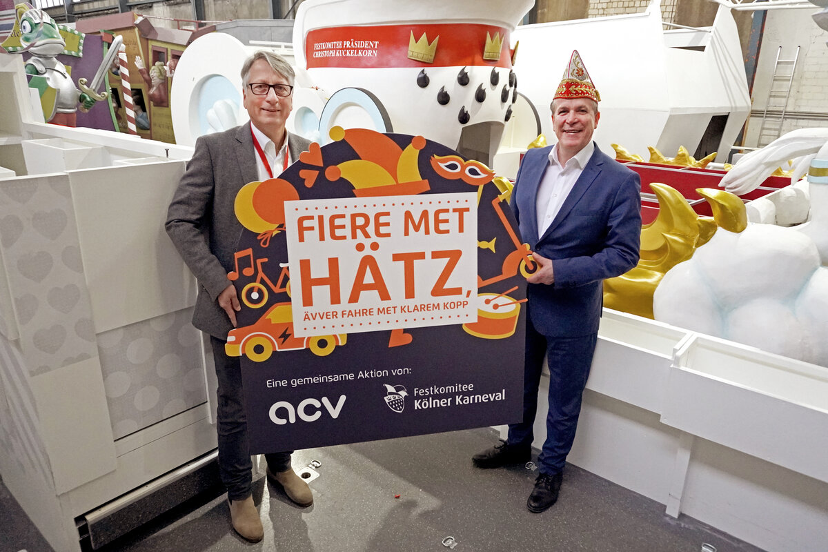 Holger Küster und Philipp Mathey halten ein großes Plakat mit der Aufschrift „Fiere met Hätz“ vor bunten Karnevalswagen.
