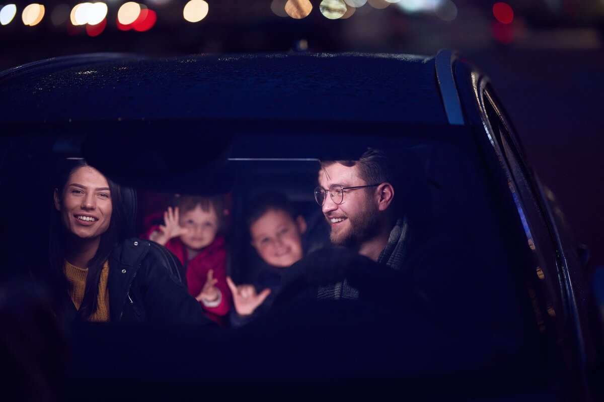 Ein Mann und eine Frau sitzen lächelnd auf den Vordersitzen eines Autos, während zwei Kinder auf dem Rücksitz in der Nacht durch die Windschutzscheibe winken und lächeln. Im verschwommenen Hintergrund sind helle Lichter zu sehen.