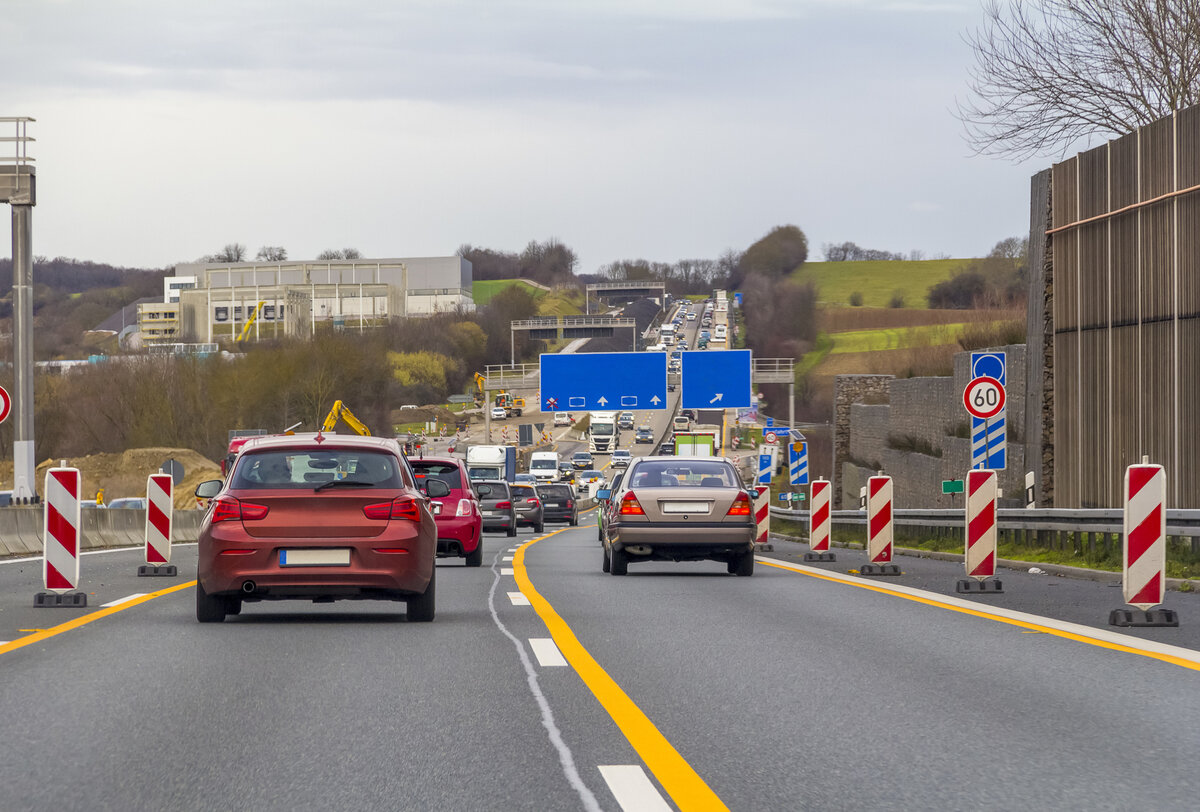 Autos fahren auf einer Autobahn mit Baustellenabsperrungen und Verkehrsschildern. Vor uns erscheinen blaue Straßenschilder, und die Straße verengt sich aufgrund der laufenden Bauarbeiten. Im Hintergrund sind Landschaften und Gebäude zu sehen.