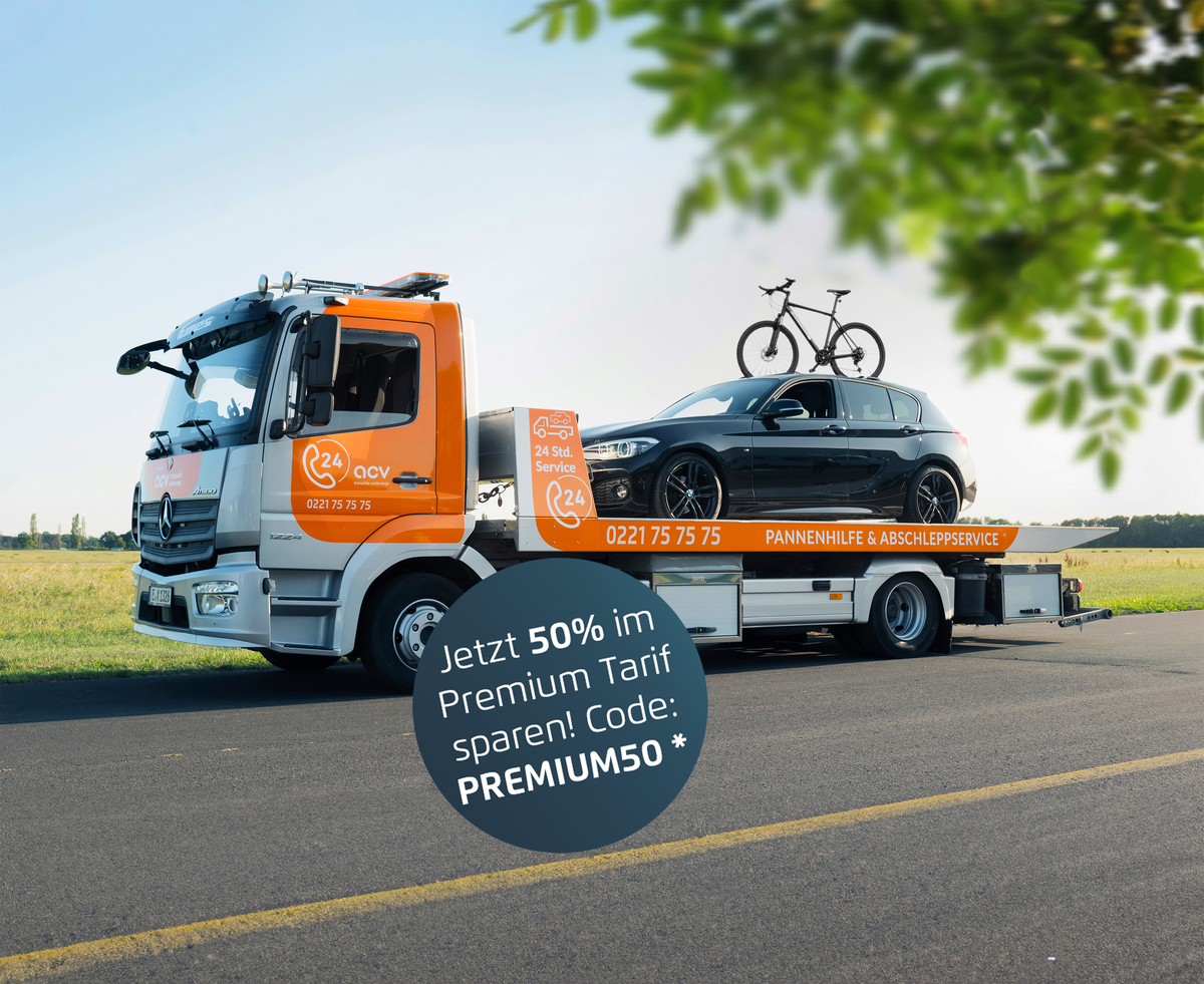 Ein orangefarbener Abschleppwagen transportiert ein schwarzes Auto mit zwei Fahrrädern auf dem Dachträger. Das Fahrzeug steht auf einer Landstraße mit Wiese und blauem Himmel im Hintergrund. Ein paar Blätter ragen von oben ins Bild.