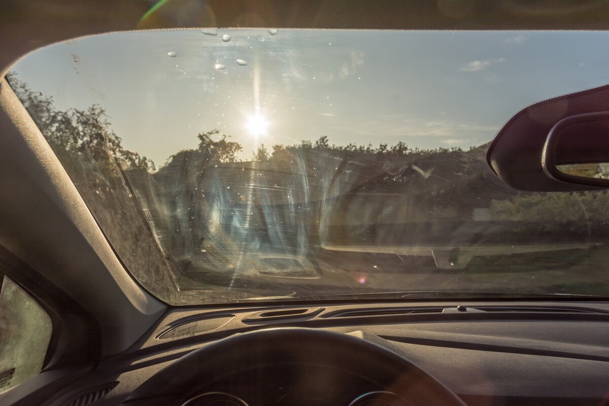 Blick aus dem Inneren eines Autos durch eine verschmutzte Windschutzscheibe mit Streifen und Flecken; draußen scheint helles Sonnenlicht und man sieht Bäume.