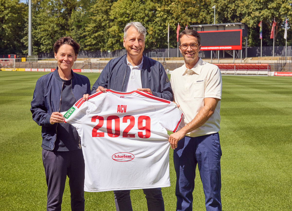 Drei Personen stehen auf einem Fußballplatz und halten ein weißes Trikot mit der Aufschrift „ACV 2029“ in den Händen. Im Hintergrund sind die Stadionränge, die Anzeigetafel und das grüne Gras zu sehen.