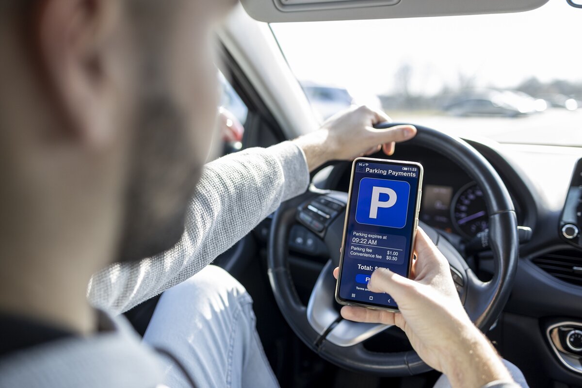 Eine Person, die in einem Auto sitzt, nutzt eine Smartphone-App, um die Parkgebühr zu bezahlen. Auf dem Bildschirm der App werden die Parkdaten angezeigt, während die Person mit einer Hand das Lenkrad und mit der anderen das Smartphone hält.