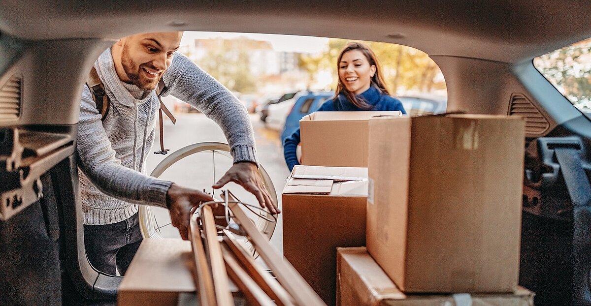 Paar lädt Umzugskartons und lange Holzlatten in den Kofferraum eines Autos, während sie gemeinsam den Umzug vorbereiten.