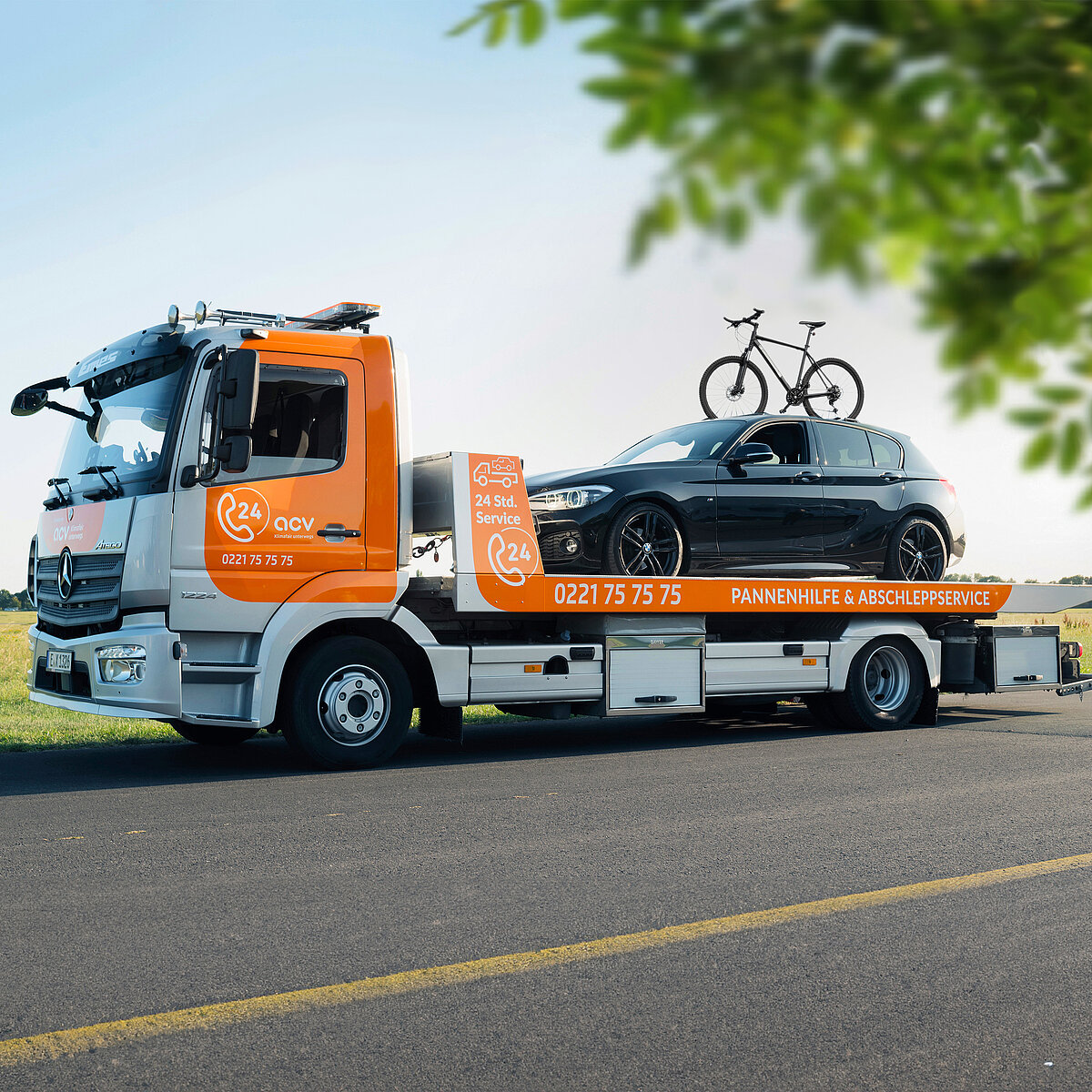 Ein orangefarbener Abschleppwagen transportiert ein schwarzes Auto mit zwei Fahrrädern auf dem Dachträger. Das Fahrzeug steht auf einer Landstraße neben einer grünen Wiese unter blauem Himmel. Im Vordergrund verläuft eine gelbe Straßenmarkierung, und ein grüner Ast ragt von oben ins Bild.