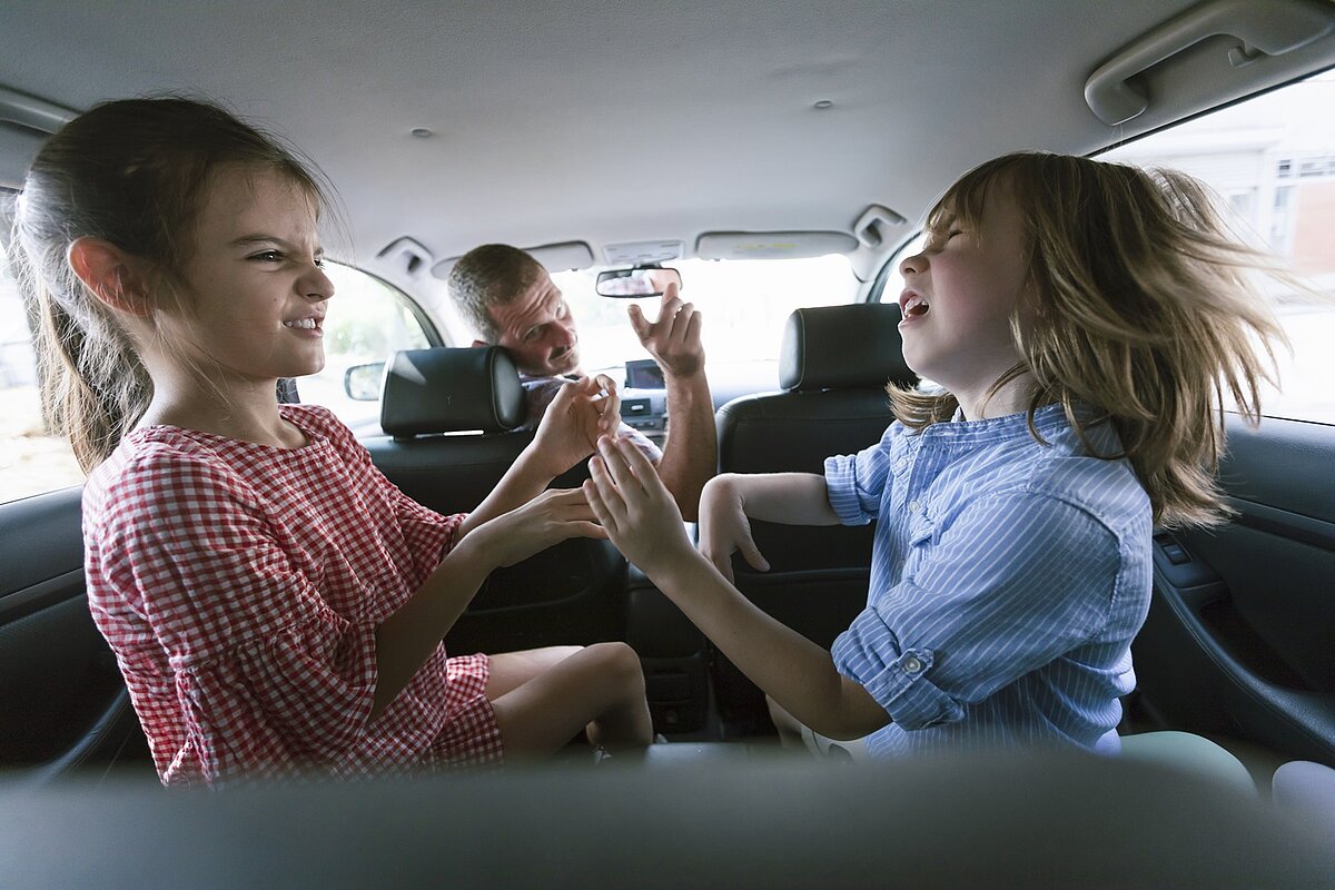 Zwei Kinder streiten sich auf der Rückbank eines Autos, während der Vater sie vom Fahrersitz aus ermahnt.