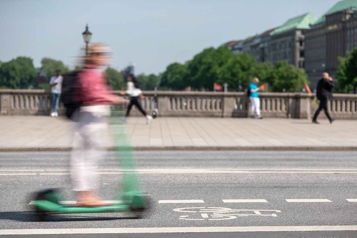 Eine unscharf abgebildete Person fährt mit einem Elektroroller auf einer Stadtstraße, auf der ein Fahrradstreifensymbol zu sehen ist, während im Hintergrund Menschen in der Nähe einer Steinbrüstung und von Bäumen stehen und gehen.