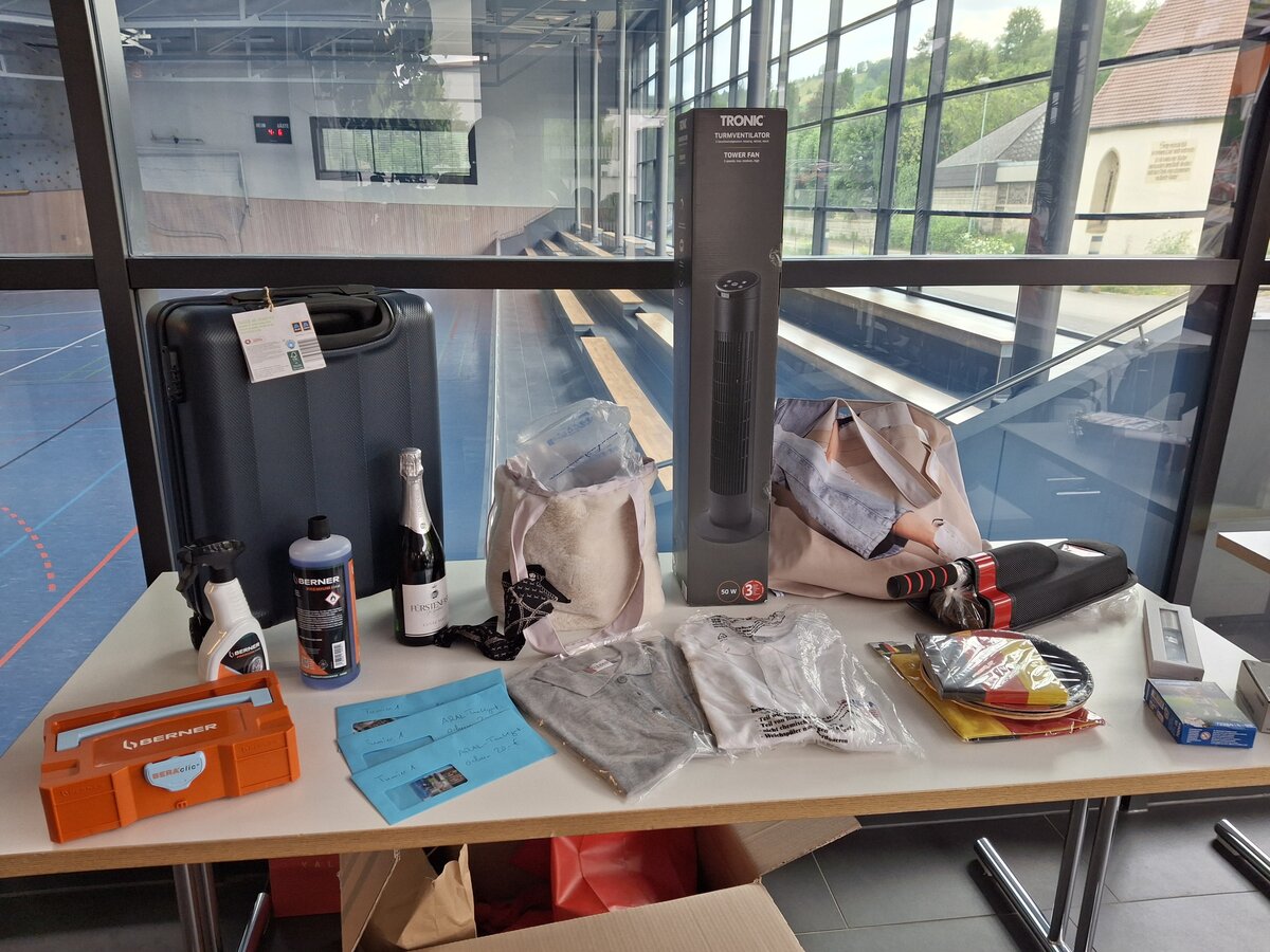 Auf einem Tisch liegen verschiedene Gegenstände, darunter ein Koffer, ein Ventilator, eine Bohrmaschine, eine Champagnerflasche, Reinigungsmittel, Kleidung, ein Werkzeugkasten, eine Tragetasche und weitere verschiedene Preise. Im Hintergrund sind ein Fitnessraum und große Fenster zu sehen.