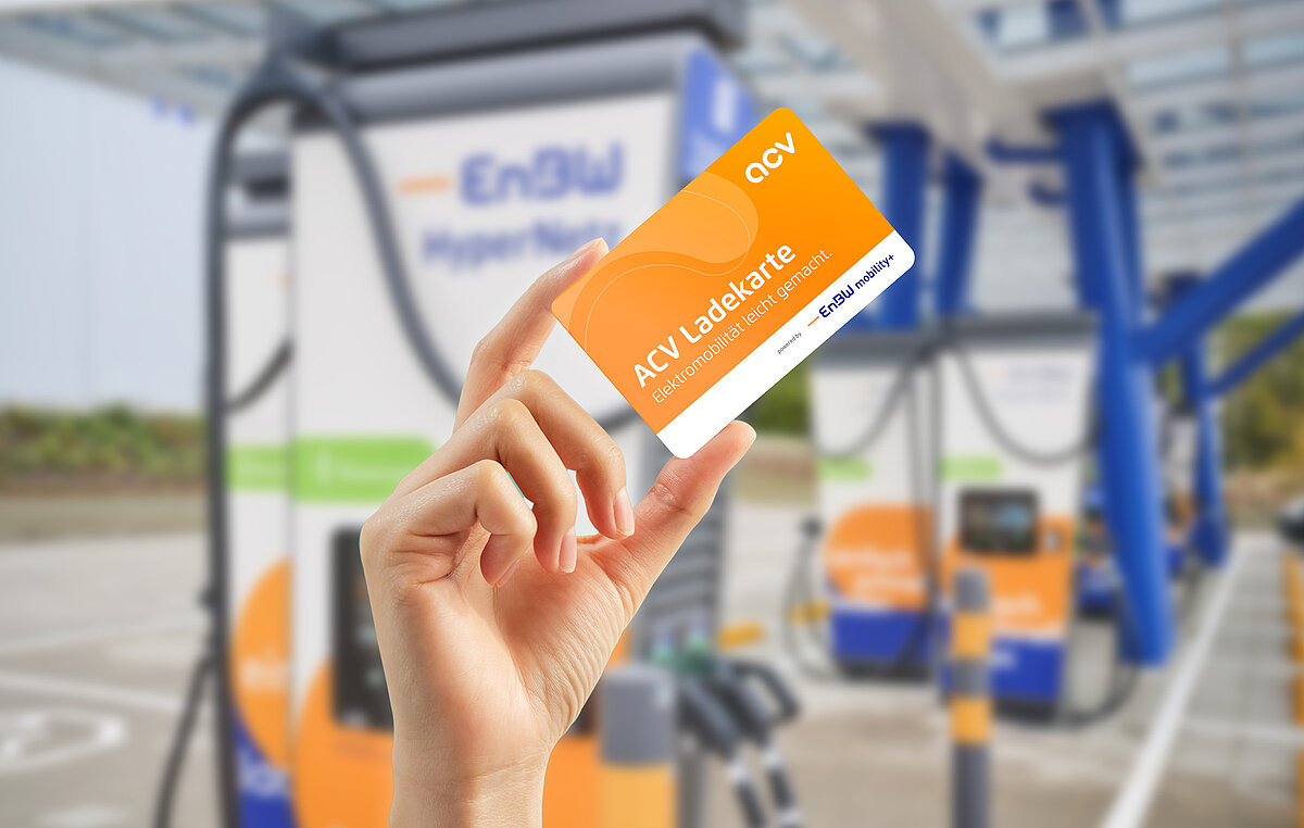 Eine Hand hält eine orange-weiße ACV-Ladekarte an einer Ladestation für Elektrofahrzeuge hoch. Im Hintergrund sind das EnBW-Logo und die Ladegeräte zu sehen.