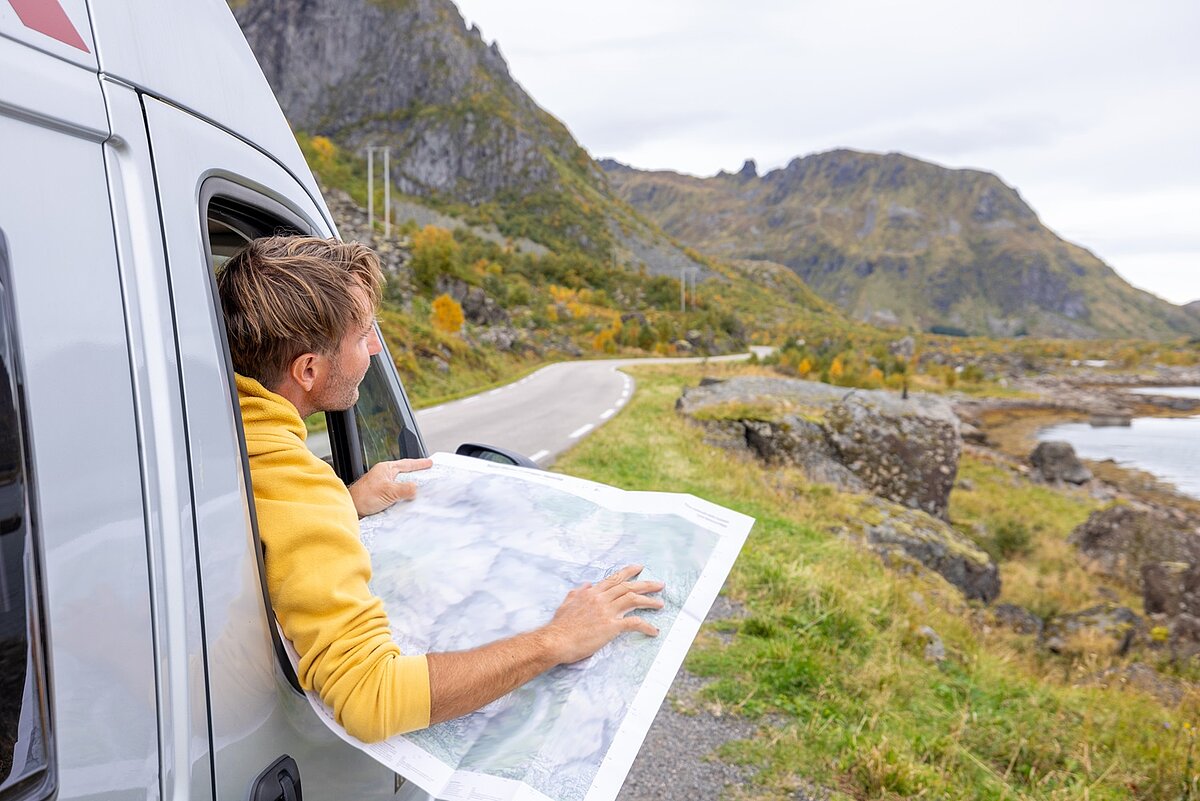 Person schaut im Camper auf eine Landkarte und plant die Route entlang einer kurvigen Straße in bergiger Landschaft.