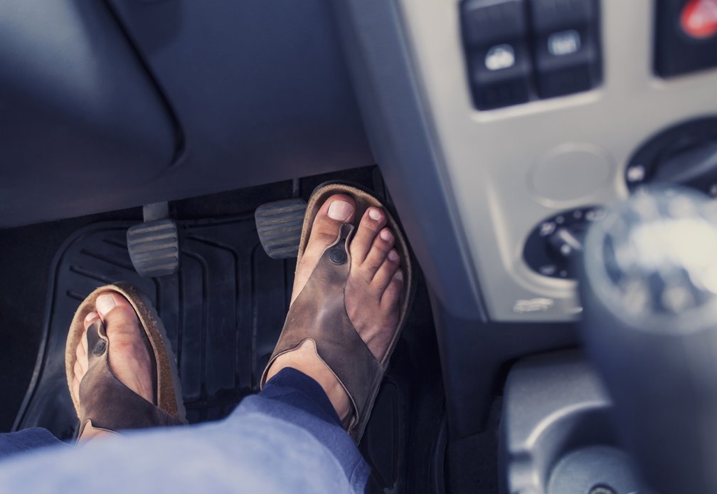Fahrer sitzt am Steuer eines Autos und trägt braune Sandalen. Der rechte Fuß ruht auf dem Gaspedal, der linke Fuß auf dem Boden im Fußraum.