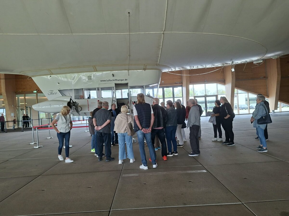 Eine Gruppe von Menschen steht unter einem großen weißen Luftschiff in einem geräumigen Hangar und hört einem Führer zu. Das Luftschiff schwebt über ihnen, und der Hangar verfügt über große Fenster und Holzbalken.