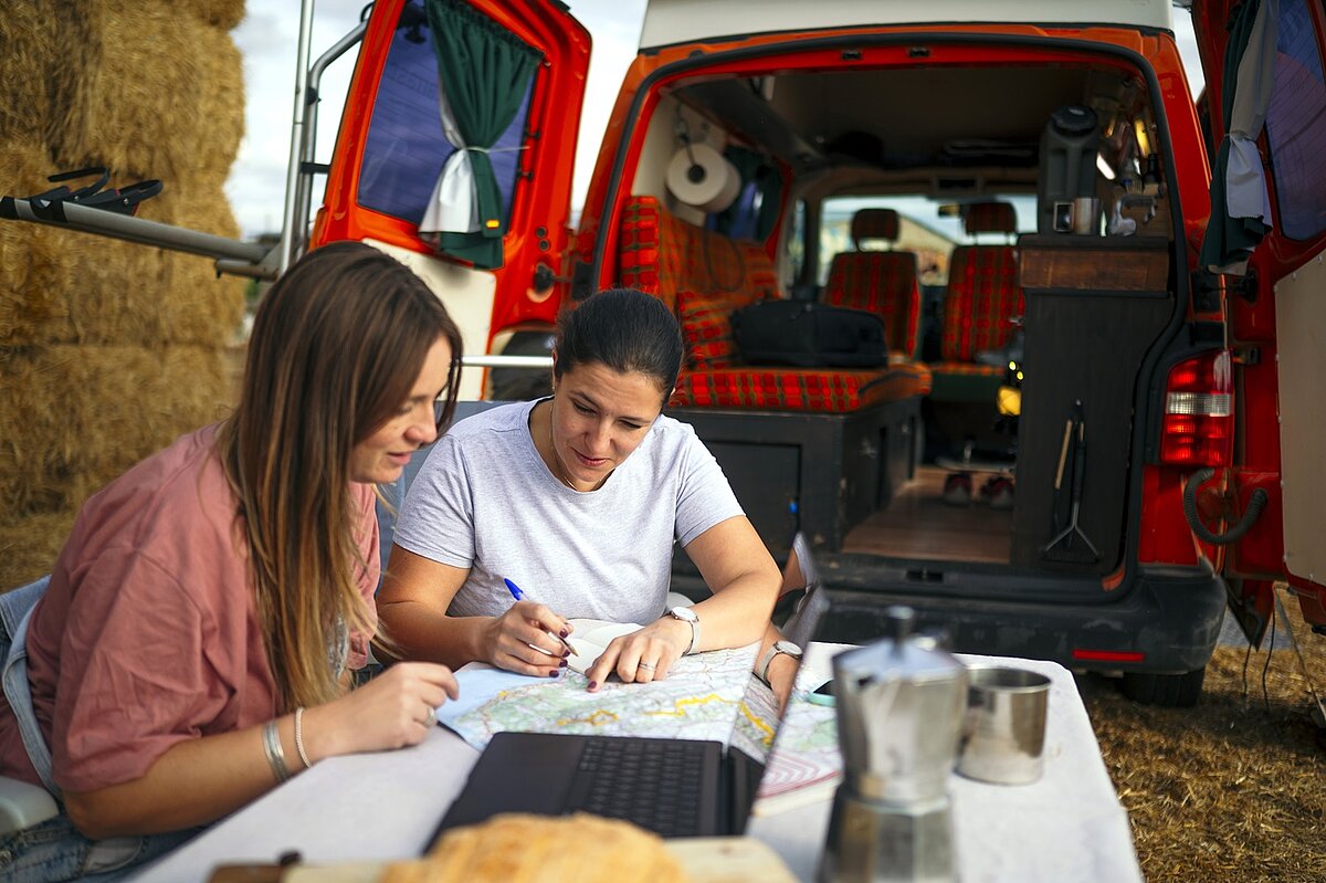 Zwei Personen sitzen vor einem Camper und planen mit Karte und Laptop ihre Reiseroute, im Hintergrund ist der geöffnete Camper mit Campingausstattung zu sehen.