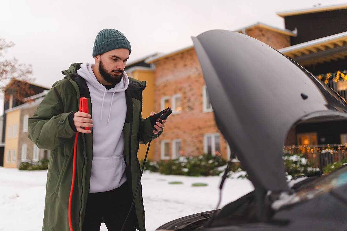 Junger Mann steht im Winter vor seinem Auto mit offenem Motorraum.