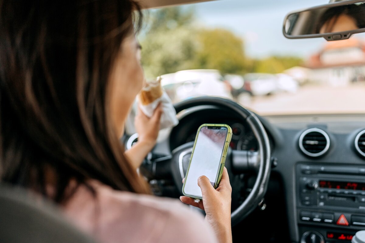 Eine Frau sitzt auf dem Fahrersitz eines Autos, hält in der einen Hand ein Sandwich und in der anderen ein Smartphone, wobei das Auto geparkt ist und das Armaturenbrett zu sehen ist.