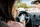 Eine Frau sitzt auf dem Fahrersitz eines Autos, hält in der einen Hand ein Sandwich und in der anderen ein Smartphone, wobei das Auto geparkt ist und das Armaturenbrett zu sehen ist.