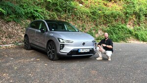 Ein Mann, der kniet und auf einen silbernen Volvo-Elektro-SUV zeigt, der auf dem Asphalt geparkt ist.
