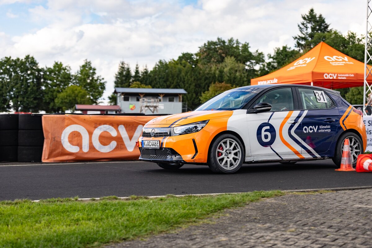 Ein weiß-orangefarbener Rennwagen mit der Startnummer 6 steht auf einer Rennstrecke in der Nähe von orangefarbenen ACV-Bannern und einem Zelt, im Hintergrund sind Bäume und ein teilweise bewölkter Himmel zu sehen.