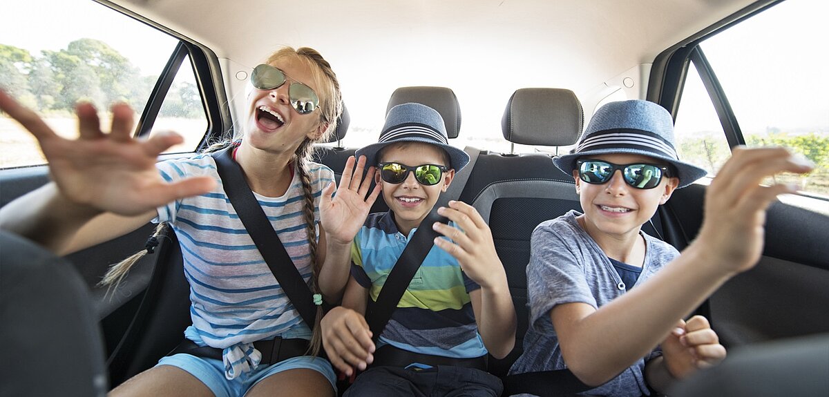 Drei fröhliche Kinder sitzen auf der Rückbank eines Autos, tragen Sonnenbrillen und Sommerkleidung, lachen und winken in die Kamera während einer Autofahrt an einem sonnigen Tag.