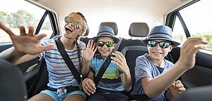 Drei fröhliche Kinder sitzen auf der Rückbank eines Autos, tragen Sonnenbrillen und Sommerkleidung, lachen und winken in die Kamera während einer Autofahrt an einem sonnigen Tag.