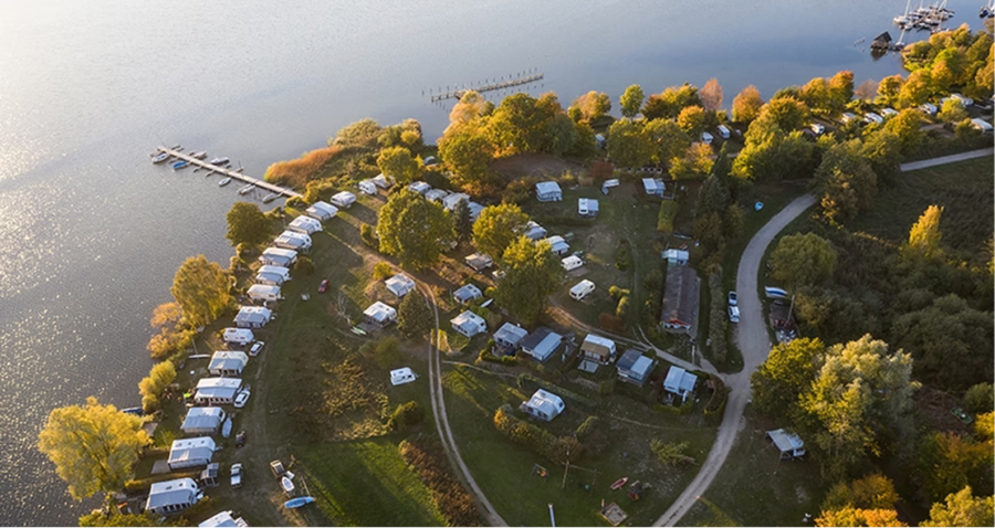 Luftaufnahme eines Campingplatzes am See mit Reihen von Wohnwagen und Zelten zwischen Bäumen, kurvenreichen Straßen und einem Steg, der in der oberen linken Ecke ins Wasser ragt. Die Szene ist in warmes Sonnenlicht getaucht.