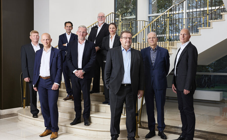 Zehn Personen in Geschäftskleidung stehen auf Marmorstufen und -böden in einer hell beleuchteten Lobby mit großen Fenstern und goldenen Geländern und posieren für ein formelles Gruppenfoto.