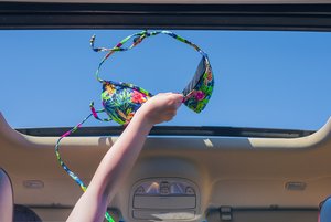 Eine Person hält einen bunten Bikini-Oberteil aus dem offenen Schiebedach eines Autos in den blauen Himmel.