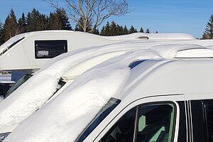 Mehrere Wohnmobile stehen nebeneinander und sind mit einer dicken Schneeschicht bedeckt, im Hintergrund Bäume und blauer Winterhimmel.