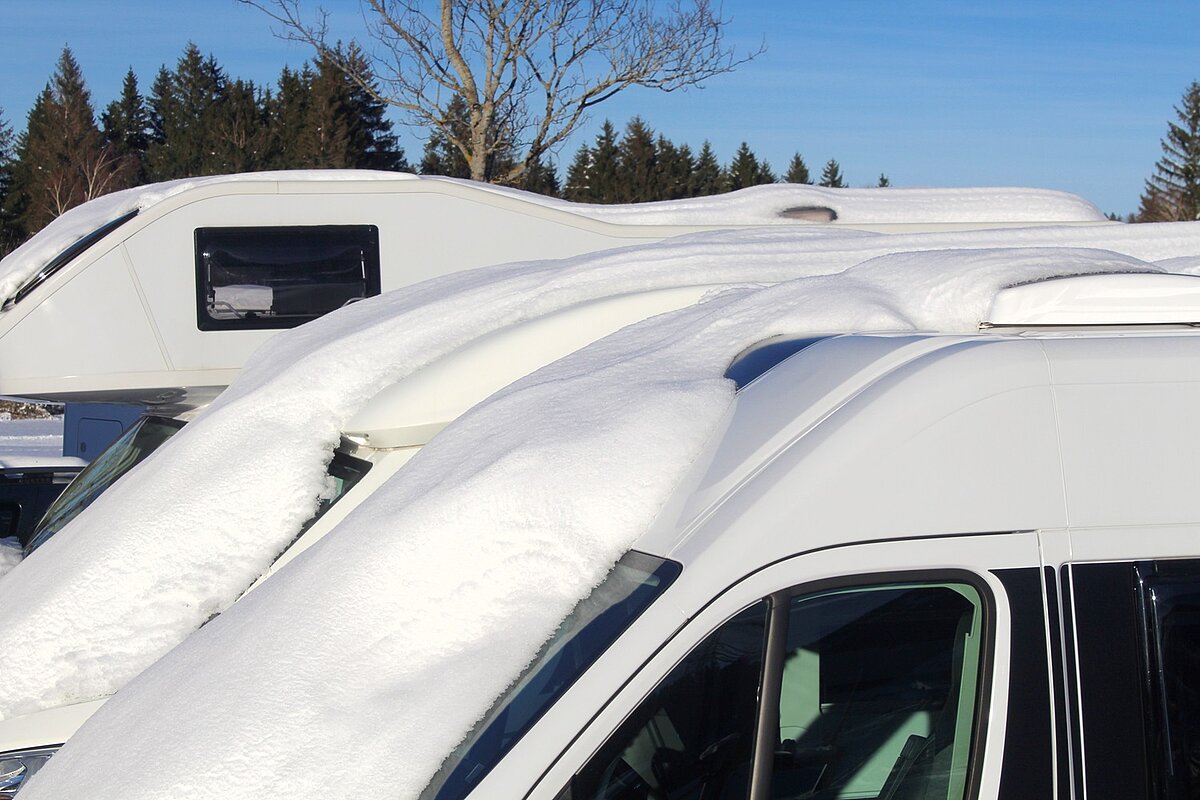 Mehrere Wohnmobile stehen nebeneinander und sind mit einer dicken Schneeschicht bedeckt, im Hintergrund Bäume und blauer Winterhimmel.