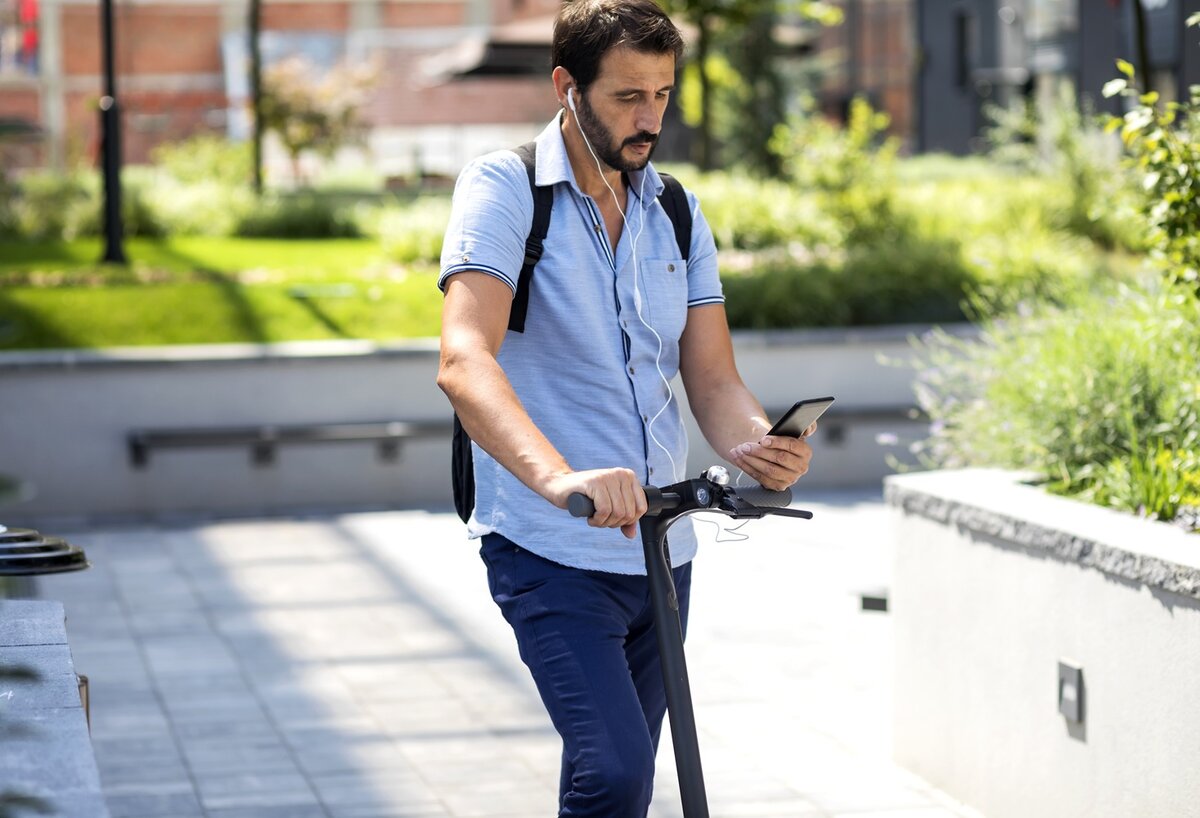 Ein Mann mit Kopfhörern und Rucksack fährt draußen auf einem Elektroroller und schaut dabei auf sein Smartphone. Die Szene ist sonnig, im Hintergrund sind Grünflächen und Gebäude zu sehen.