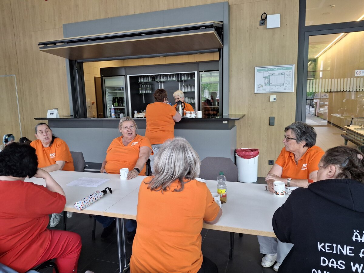 Eine Gruppe von Frauen, die größtenteils orangefarbene T-Shirts tragen, sitzt an einem Tisch neben einer Bar. Die Frau links hat blaue Haare und trägt Kopfhörer.