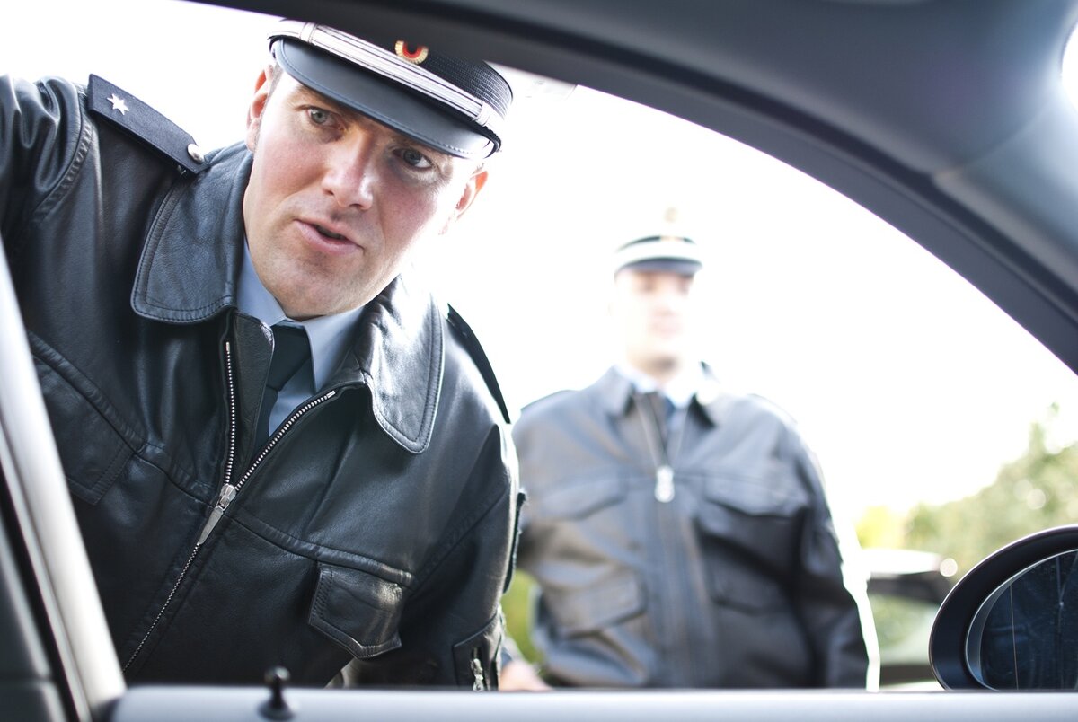 Ein Polizeibeamter in Uniform beugt sich hinunter, um in ein Autofenster zu schauen, während ein anderer Beamter im Hintergrund außerhalb des Fahrzeugs steht. Die Szene scheint eine Verkehrskontrolle zu sein.