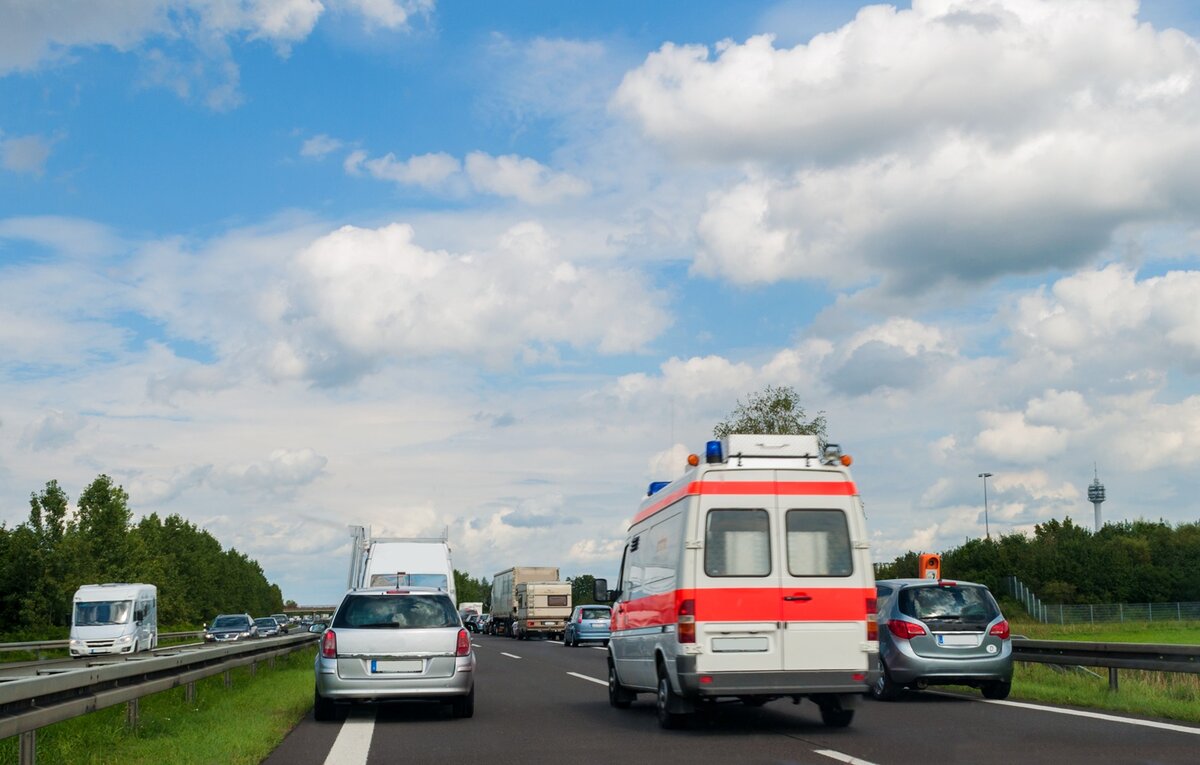 Ein Krankenwagen mit Blaulicht fährt auf einer Autobahn und überholt unter einem teilweise bewölkten Himmel Autos und andere Fahrzeuge. Im Hintergrund säumen Bäume die Straße.