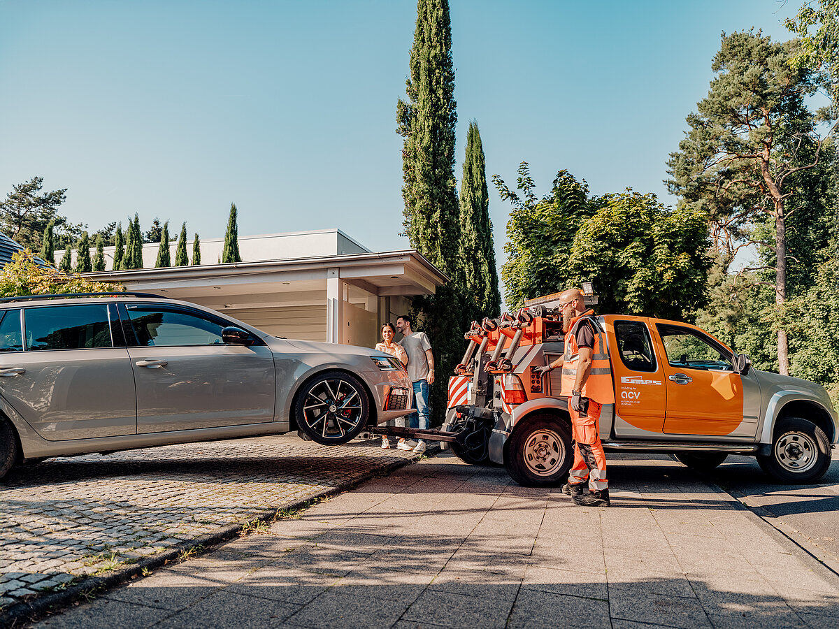 „Pannenhelfer lädt ein silbernes Auto mit angehobenen Vorderrädern auf einen orangefarbenen Abschleppwagen, während ein Paar zusieht.“