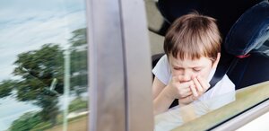 Ein kleiner Junge sitzt auf einem Autositz am Fenster, hat die Augen geschlossen und hält sich mit beiden Händen den Mund zu, weil er sich während einer Autofahrt tagsüber unwohl zu fühlen scheint.
