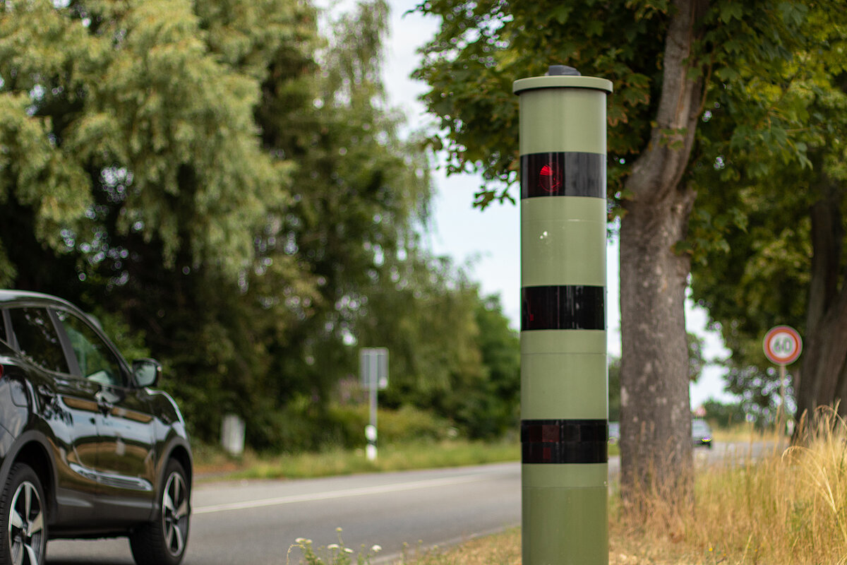 schwarzes Auto fährt an einem Blitzer auf der rechten Straßenseite vorbei