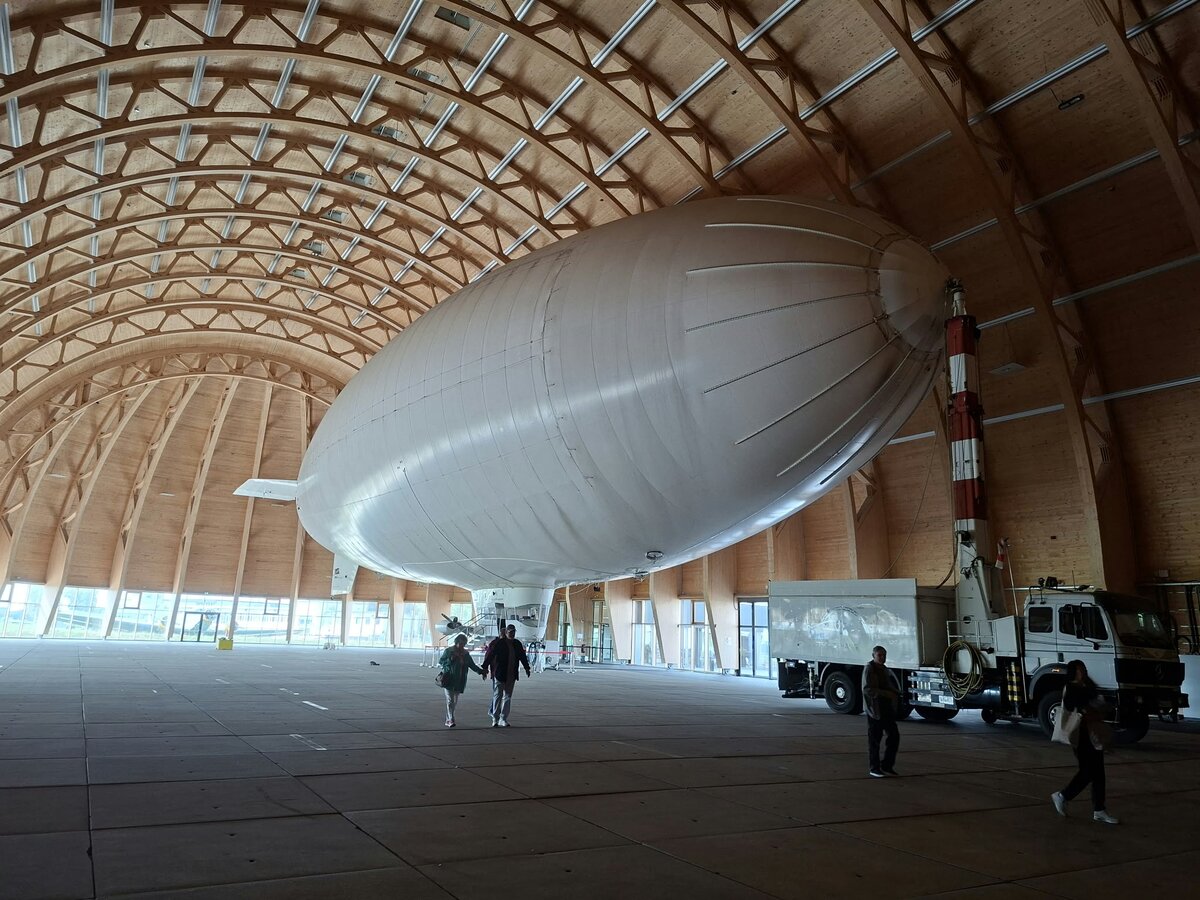 Ein großes Luftschiff steht in einem geräumigen, gewölbten Holzhangar. In der Nähe befinden sich mehrere Personen und ein Lastwagen, was die immense Größe des Luftschiffs und des Gebäudes noch unterstreicht.