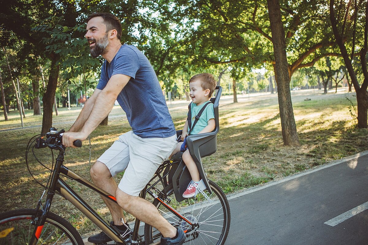 Vater fährt mit Kind im Fahrradsitz auf einem Radweg im Park.