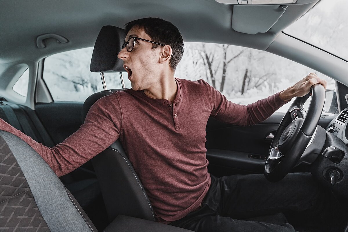 Junger Mann mit Brille und rotem Langarmshirt sitzt am Steuer eines Autos, dreht sich nach hinten und blickt konzentriert über die rechte Schulter – typische Szene beim Einparken.