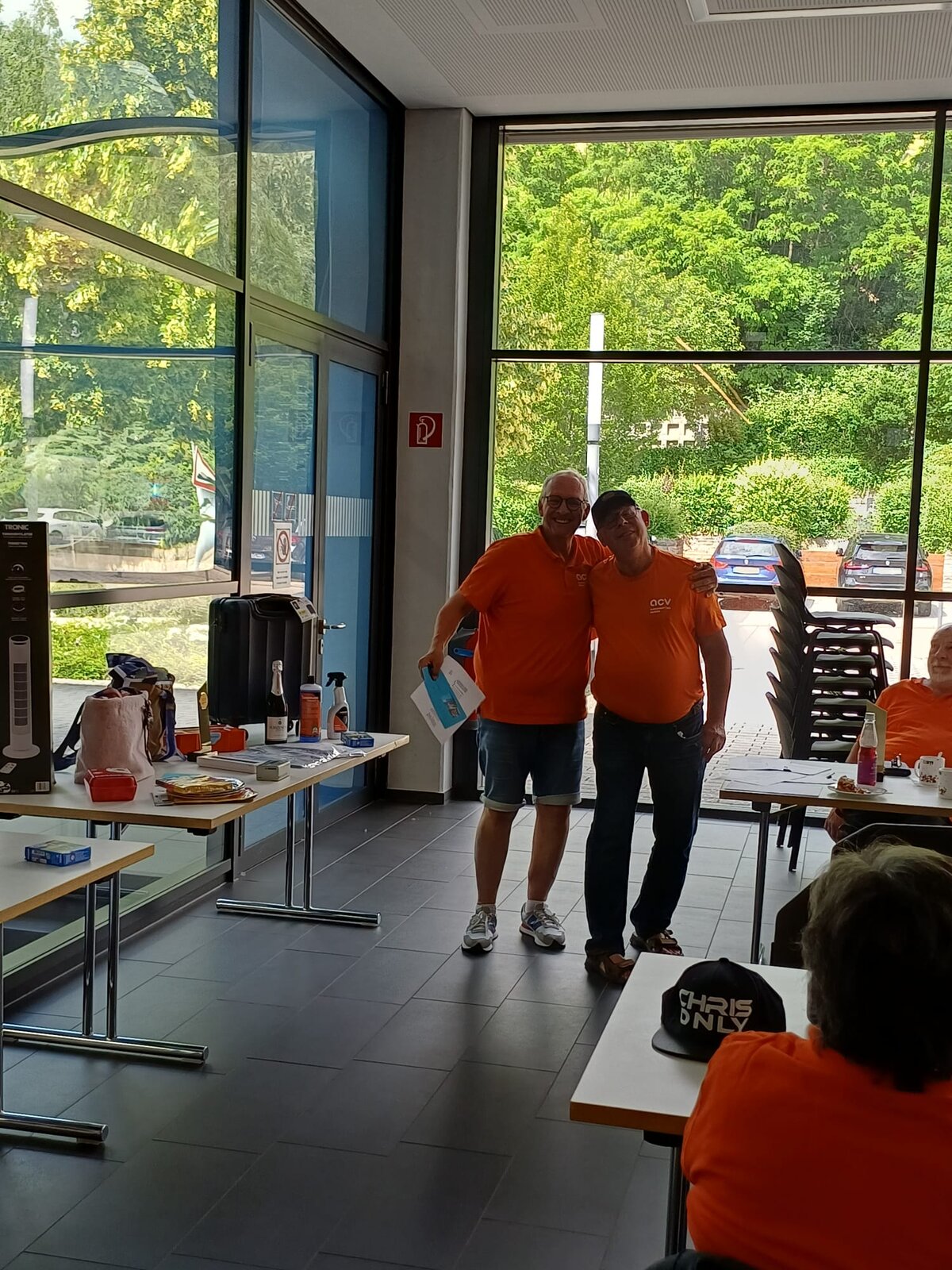 Zwei Männer in orangefarbenen Hemden stehen lächelnd nebeneinander. Der eine hat seinen Arm um die Schultern des anderen gelegt. Im Hintergrund steht ein Tisch, der mit Geschenken und Auszeichnungen beladen ist.