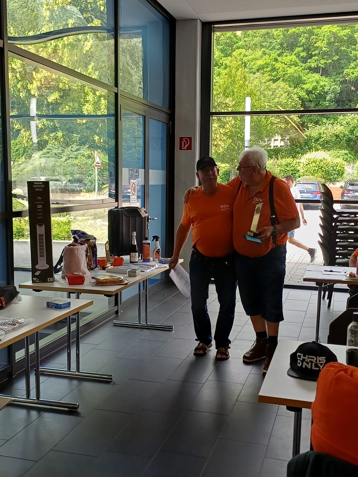 Zwei Männer in identischen orangefarbenen ACV-Shirts stehen bei einer Veranstaltung gemeinsam in einem Raum, einer von ihnen hält einen Pokal in der Hand. Zu sehen sind Tische mit Preisen und Fanartikeln sowie große Fenster mit Blick auf eine begrünte Außenanlage.
