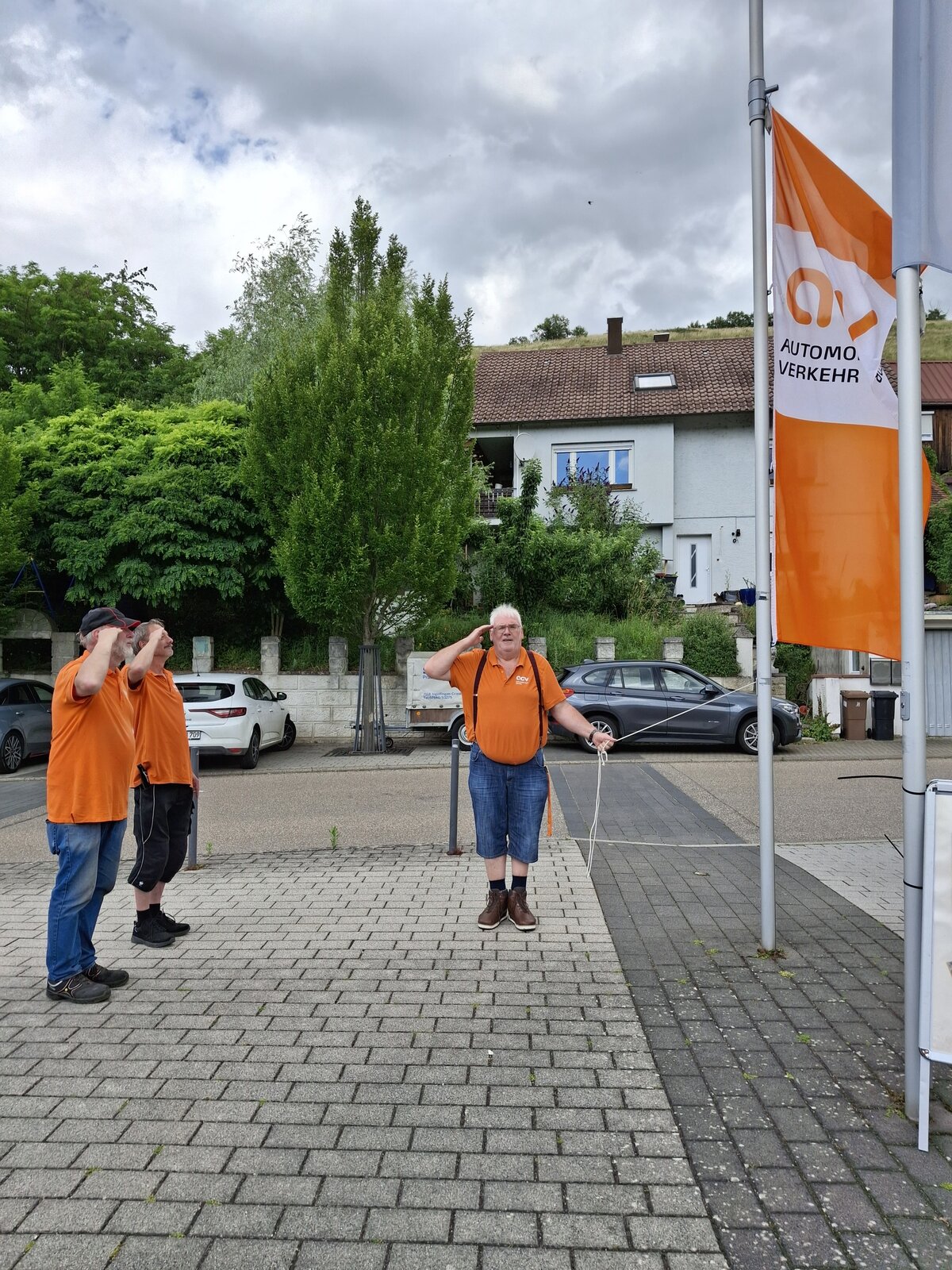 Drei Personen in orangefarbenen Hemden salutieren vor einer orangefarbenen „Automobil-Verkehr“-Flagge an einem Mast im Freien, im Hintergrund ist eine Wohnstraße mit Häusern und Grünflächen zu sehen.