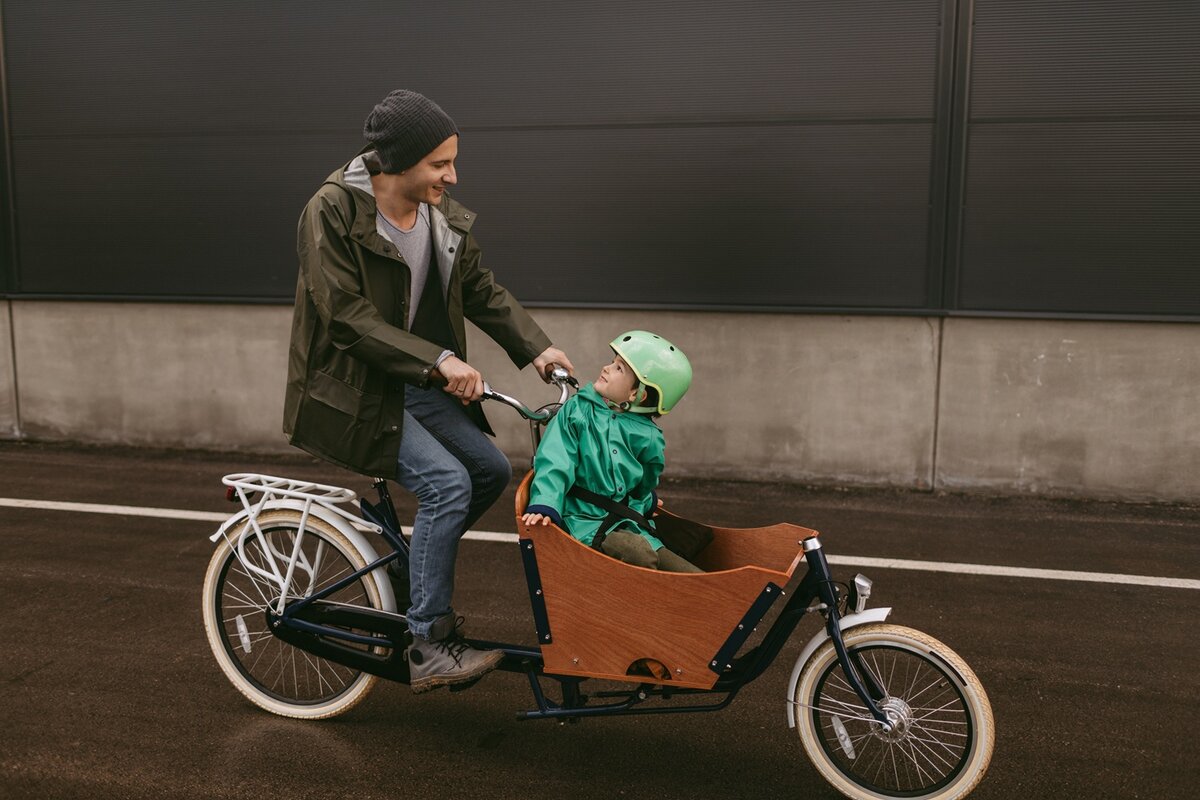 Ein Erwachsener fährt auf einem Lastenrad, auf dem ein kleines Kind im vorderen Holzfach sitzt. Beide tragen Jacken, und das Kind hat einen grünen Helm auf. Sie lächeln sich auf einer gepflasterten Fläche an.