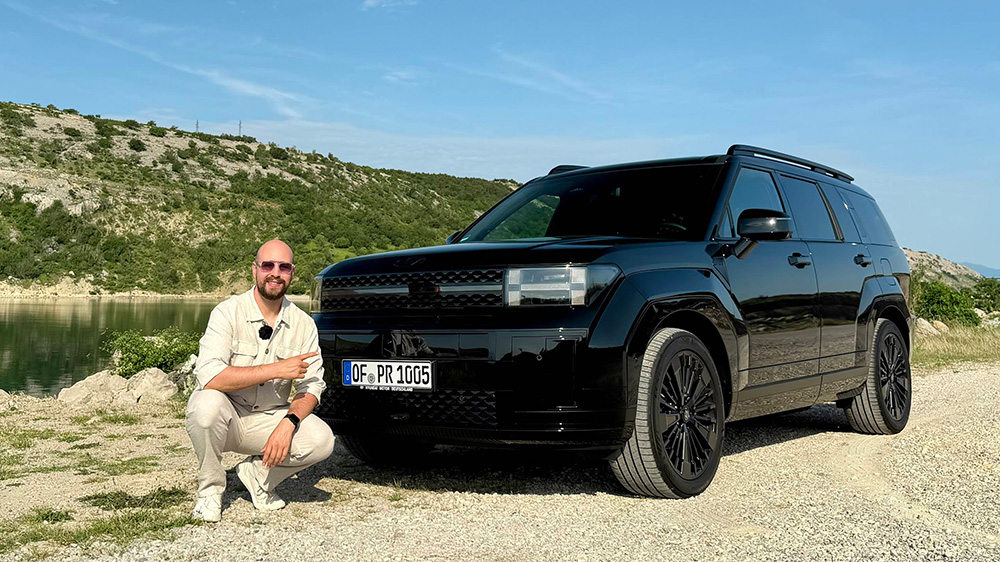 Eine Person in leichter Kleidung kniet lächelnd neben einem glänzenden schwarzen SUV, der auf einer Schotterstraße geparkt ist, im Hintergrund sind Grünflächen und Hügel vor einem blauen Himmel zu sehen.