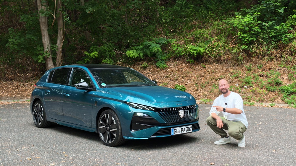 Ein Mann hockt neben einem blauen Fließheckwagen, der auf einer gepflasterten Fläche geparkt ist; im Hintergrund sind grüne Bäume und Büsche zu sehen.