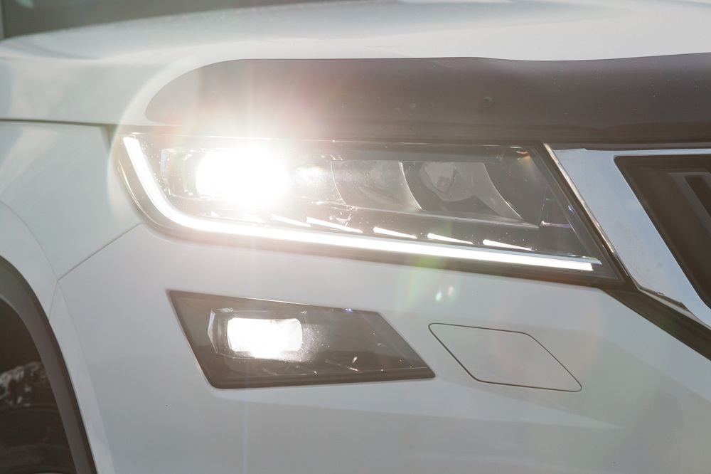 Das Bild zeigt einen Nahaufnahmeausschnitt des Frontscheinwerfers eines weißen Autos, das bei strahlendem Sonnenschein fotografiert wurde. Das Tagesfahrtlicht ist eingeschaltet, und das helle Licht strahlt direkt in die Kamera.