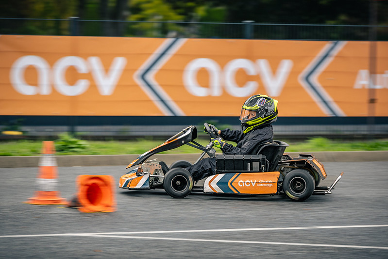 Eine Person in einem schwarzen Rennanzug und mit Helm fährt mit einem orangefarbenen Go-Kart an orangefarbenen Verkehrskegeln vorbei auf einer gepflasterten Strecke, im Hintergrund sind ACV-Banner zu sehen.