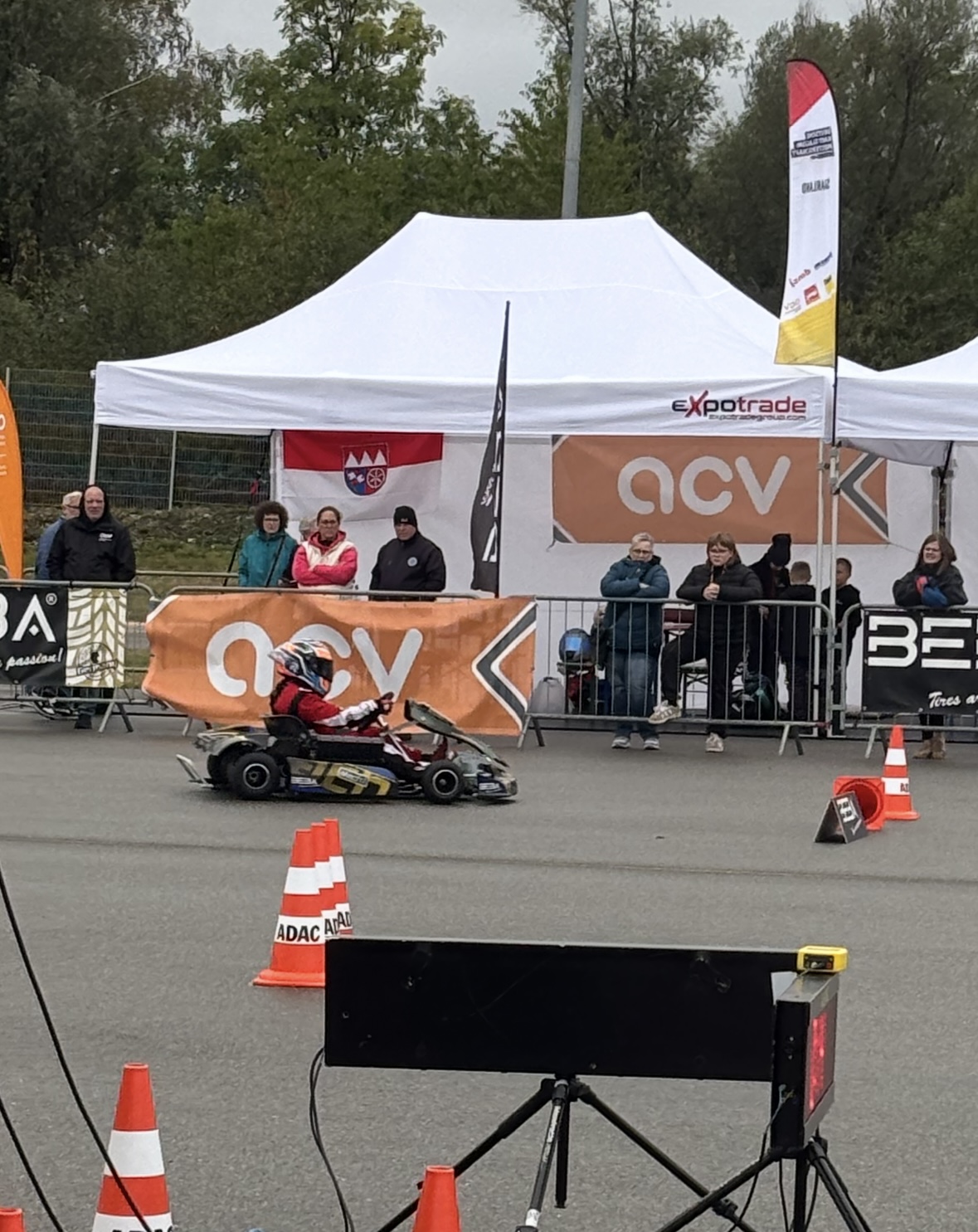Ein Kind in Rennanzug und Helm fährt vor Zuschauern und weißen Veranstaltungszelten auf einer Asphaltstrecke Go-Kart.